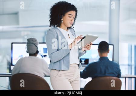 Junge Geschäftsfrau mit gemischter Rasse, die auf einem digitalen Tablet-Gerät surft, während sie in einem Callcenter mit ihren Kollegen im Hintergrund arbeitet. Ernst Stockfoto