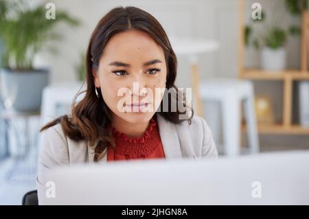 Junge selbstbewusste asiatische Geschäftsfrau, die allein in einem Büro sitzt und am Computer im Internet surft. Ambitionierter Kreativprofi Stockfoto
