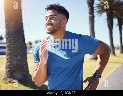 Glücklich lächelnder junger, gemischter Mann, der allein im Sommer einen Lauf in einem Park genießt. Ein fröhlicher hispanischer Mann lächelt, trainiert und joggt Stockfoto