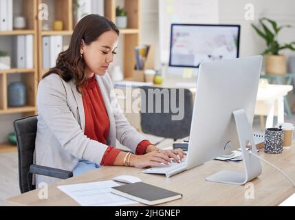 Junge, selbstbewusste asiatische Geschäftsfrau, die allein in einem Büro sitzt und am Computer im Internet surft. Ambitioniertes kreatives, professionelles Networking Stockfoto