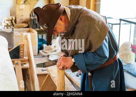 Ein älterer Mann, der in einem mittelalterlichen Kostüm aus einer blauen Tunika und einem braunen Filzhut gekleidet ist, beugt sich über einen Holzstab, während er ihn in einem mittelalterlichen Arbeitszimmer einordnen wird. Stockfoto