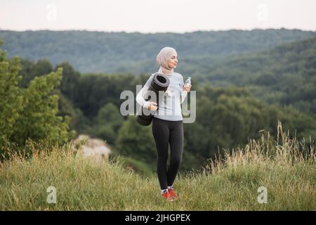 Sportliche, gesunde Frau im Hijab, die Yogamatte und eine Flasche Wasser hält, während sie im Stadtpark steht. Lächelnde muslimische Frau, die während des Trainings im Freien Pause nimmt. Stockfoto