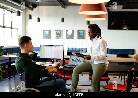 Der junge afroamerikanische Geschäftsmann diskutiert mit einem kaukasischen Kollegen, der im Rollstuhl sitzt Stockfoto