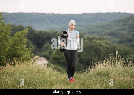 Sportliche, gesunde Frau im Hijab, die Yogamatte und eine Flasche Wasser hält, während sie im Stadtpark steht. Lächelnde muslimische Frau, die während des Trainings im Freien Pause nimmt. Stockfoto