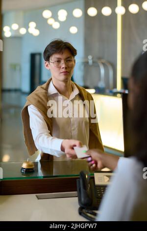 Junger asiatischer Mann in Casualwear, der seinen Pass an den Rezeptionisten über den Schalter übergab, während er bei der Ankunft im Hotel die Registrierung abnahm Stockfoto