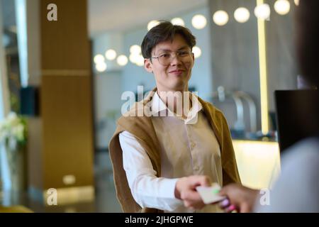 Fröhlicher junger Tourist in eleganter Freizeitkleidung und Brillen, der die Schlüsselkarte an die Rezeption übergibt oder sie nach der Registrierung im Hotel abnimmt Stockfoto