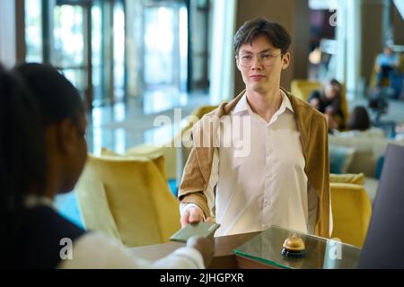 Ein junger seriöser Geschäftsmann asiatischer Herkunft übergibt seinen Pass an der Rezeption am Schalter während der Registrierung im luxuriösen Hotel Stockfoto