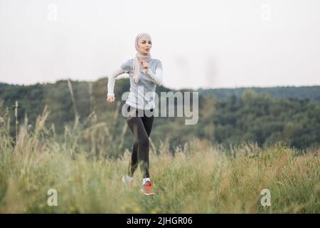 Glückliche arabische Frau in Hijab in aktiven Rennen im Sommerpark. Junge Frau, die ihre Freizeit an der frischen Luft verbringt. Lächelnde muslimische Frau beim Joggen im Freien. Stockfoto