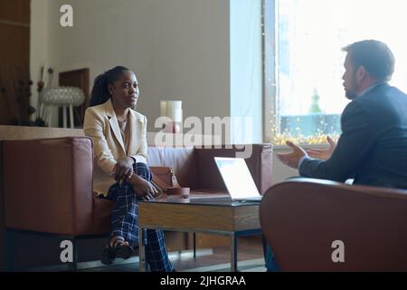 Junge elegante schwarze Frau, die beim Treffen auf den kaukasischen männlichen Geschäftspartner schaut und ihre Arbeitspunkte oder Finanzdaten erklärt Stockfoto