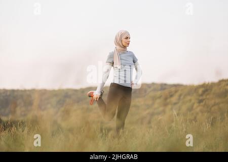 Aktive Frau in Sportkleidung und Hijab Stretching Beine im Sommerpark. Junges arabisches Mädchen, das am Morgen auf dem Hintergrund eines schönen Parks steht und körperliche Übungen macht Sportliche Aktivitäten im Freien. Stockfoto