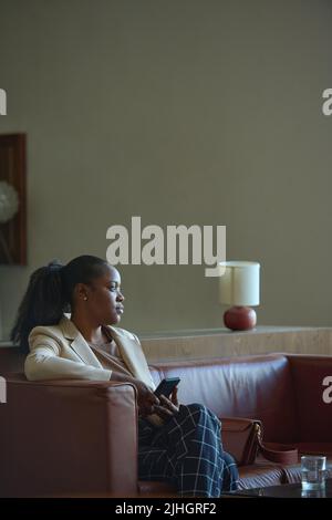 Junge erholsame afroamerikanische Geschäftsfrau in eleganter Casualwear, die auf dem Ledersofa im Hotelzimmer oder in der Lounge sitzt und das Smartphone benutzt Stockfoto