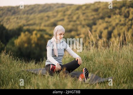 Fröhliche junge Sportlerin im Hijab und Aktivohr, die die Beinmuskeln streckt, während sie auf der Matte im Park sitzt. Porträt einer attraktiven jungen muslimischen Frau, die ihren Körper dehnt und im Freien trainiert Stockfoto