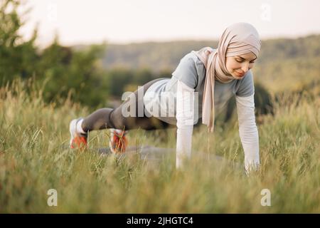Porträt einer attraktiven jungen muslimischen Frau, die lächelt und am Brett tust. Glückliche junge muslimische Frau, die im grünen Park Sport macht. Gesunder Lebensstil und gute Wellness-Konzepte. Stockfoto