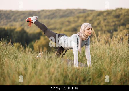 Lächelnde junge Frau im Hijab- und Sportoutfit, die während des morgendlichen Workouts an der frischen Luft flexible Übungen auf der Yogamatte macht. Training im Sommerpark. Gesunde Lebensweise. Stockfoto