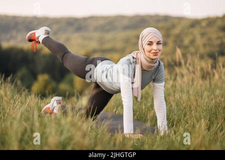 Lächelnde junge Frau im Hijab- und Sportoutfit, die während des morgendlichen Workouts an der frischen Luft flexible Übungen auf der Yogamatte macht. Training im Sommerpark. Gesunde Lebensweise. Stockfoto