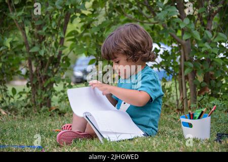 Der kleine Junge verwendet einen Zauberstift, um Bilder in einem Buch auf einem Holztisch im Park mit Gesichtsausdruck der Entschlossenheit zu zeichnen. Stockfoto