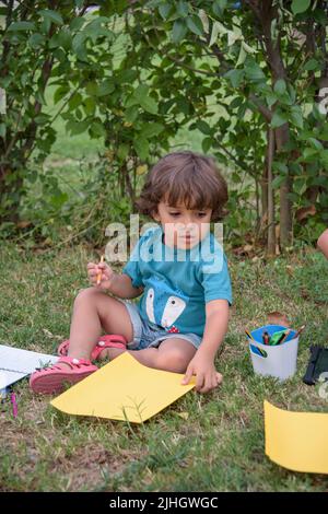 Der kleine Junge verwendet einen Zauberstift, um Bilder in einem Buch auf einem Holztisch im Park mit Gesichtsausdruck der Entschlossenheit zu zeichnen. Stockfoto