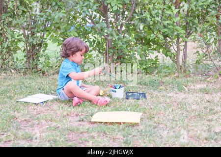 Der kleine Junge verwendet einen Zauberstift, um Bilder in einem Buch auf einem Holztisch im Park mit Gesichtsausdruck der Entschlossenheit zu zeichnen. Stockfoto