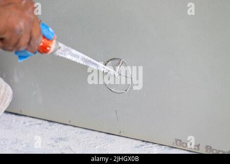 Ein Bauarbeiter schneidet mit einer Säge ein Loch für ein Rohr aus, bevor die Trockenwand auf die Rahmenstreben an der Wand montiert wird. Neu Stockfoto