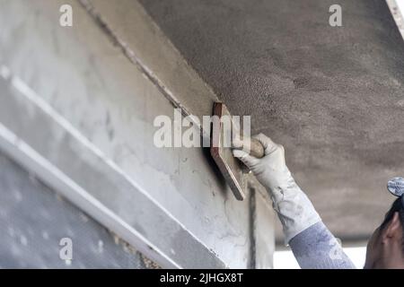 Ein gut ausgebildeter Bauarbeiter verwendet eine nasse Schwammkelle, um frisch angewendeten Stuck an der Außenwand eines Mehrfamilienhauses zu glätten. Stockfoto