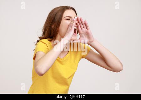 Das Profil eines Teenagers im gelben T-Shirt hält die Arme weit offen, schreit laut, macht auf wichtige Nachrichten aufmerksam und kündigt das Ereignis an. Innenaufnahme des Studios isoliert auf grauem Hintergrund. Stockfoto