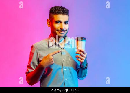 Porträt eines glücklichen optimistischen Mannes im Hemd, der den Finger auf die Tasse in der Hand zeigt und die Kamera anschaut, braucht Energie. Innenaufnahmen im Studio, isoliert auf farbigem Neonlicht-Hintergrund. Stockfoto