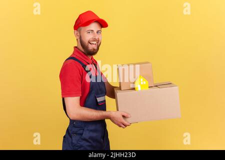 Profil Porträt des Lieferers in blauer Uniform und roter Kappe mit Pappkartons und sicherer Tür-zu-Tür-Lieferung aus dem Papierhaus. Innenaufnahme des Studios isoliert auf gelbem Hintergrund. Stockfoto