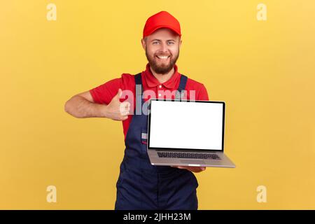 Porträt eines zufriedenen, glücklichen Arbeiters in Uniform, der seinen Laptop mit leerem Bildschirm und Werbefläche in der Hand hält und den Daumen nach oben zeigt. Innenaufnahme des Studios isoliert auf gelbem Hintergrund. Stockfoto