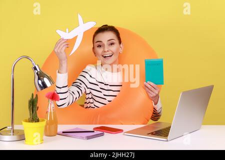 Extrem glückliche Frau, die in den Sommerferien geht, mit Gummiring und Spielzeugflugzeug im Büro sitzt und Pass zeigt, Tickets kauft. Studio-Innenaufnahme isoliert auf gelbem Hintergrund. Stockfoto