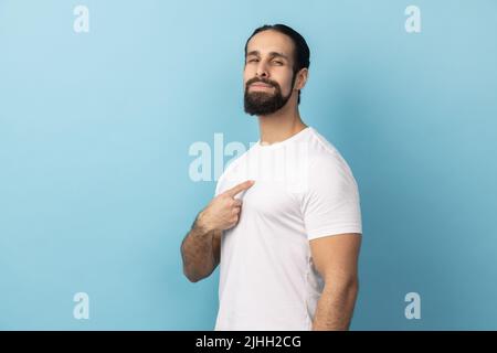 Porträt eines selbstbewussten Mannes mit weißem T-Shirt und Bart, der auf sich selbst zeigt, stolz und selbstwichtig ist und ein großes Ego hat. Innenaufnahme des Studios isoliert auf blauem Hintergrund. Stockfoto