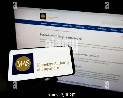 Person mit Mobiltelefon und Logo der Monetary Authority of Singapore (MAS) auf dem Bildschirm vor der Webseite. Konzentrieren Sie sich auf die Telefonanzeige. Stockfoto