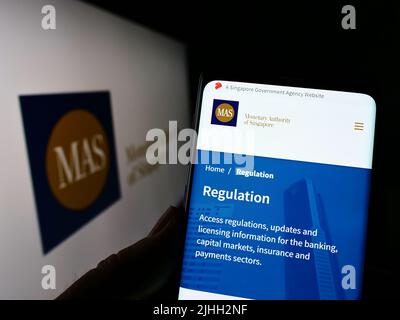Person mit Smartphone und Webseite der Monetary Authority of Singapore (MAS) auf dem Bildschirm vor dem Logo. Konzentrieren Sie sich auf die Mitte des Telefondisplays. Stockfoto
