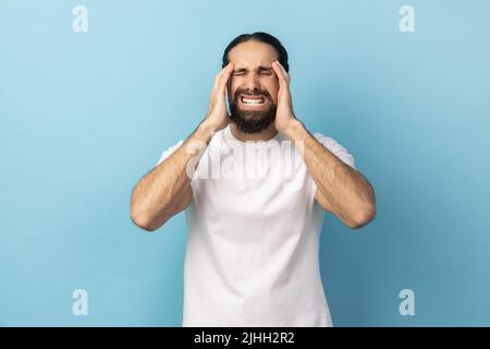 Porträt eines kranken Mannes mit Bart und weißem T-Shirt, der unter schrecklichen Kopfschmerzen litt, das Gesicht runzelte und die Finger auf den Schläfen hielt. Innenaufnahme des Studios isoliert auf blauem Hintergrund. Stockfoto