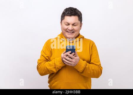 Attraktiver Mann mittleren Alters, der ein Mobiltelefon mit glücklichem Ausdruck verwendet, süchtig nach Smartphone, SMS in sozialen Netzwerken, Kapuzenpullover im urbanen Stil trägt. Innenaufnahme des Studios isoliert auf weißem Hintergrund. Stockfoto