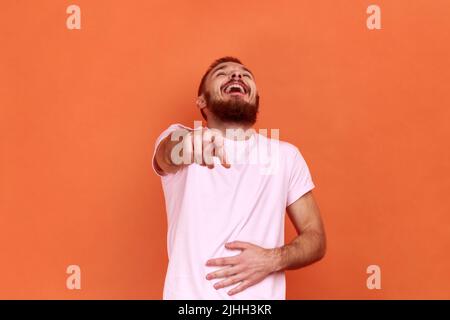 Porträt eines lustigen, positiven bärtigen Mannes, der laut lacht und den Bauch hält und mit dem Finger auf dich zeigt, Spott, in einem rosa T-Shirt. Innenaufnahme des Studios isoliert auf orangefarbenem Hintergrund. Stockfoto