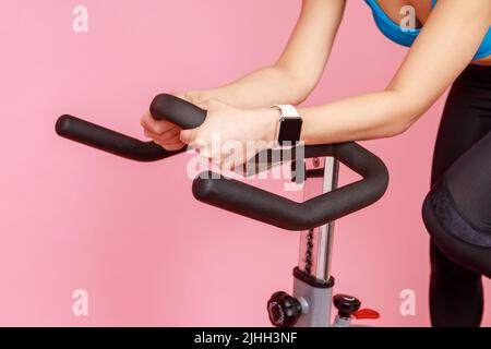 Nahaufnahme Porträt einer unbekannten Frau Hände mit Smartwatch auf Sportgerät, Reiten Heimtrainer. Innenaufnahme des Studios isoliert auf rosa Hintergrund. Stockfoto