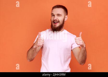 Porträt eines positiven bärtigen Mannes, der mit den Daumen nach oben steht, wie Geste, seine Zustimmung demonstriert und dem Vorschlag zustimmt, in einem rosa T-Shirt. Innenaufnahme des Studios isoliert auf orangefarbenem Hintergrund. Stockfoto