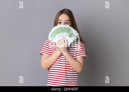 Porträt eines charmanten kleinen Mädchens in gestreiftem T-Shirt, das sich hinter einem Fan von hundert Euro-Scheinen versteckt und sich nach Geld duftet. Innenaufnahme des Studios isoliert auf grauem Hintergrund. Stockfoto