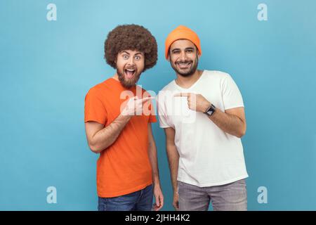 Beste Freunde. Portrait von zwei jungen erwachsenen Hipster-Männern, die sich mit dem Finger zeigen und mit positiven, fröhlichen Ausdrücken auf die Kamera blicken. Innenaufnahme des Studios isoliert auf blauem Hintergrund. Stockfoto