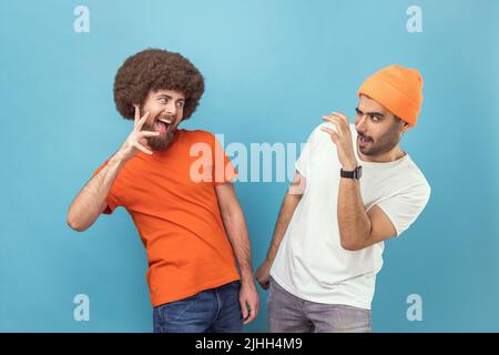 Portrait von zwei verrückten jungen erwachsenen Hipster-Männern in T-Shirts, die mit erhobenen Krallen stehen und sich mit witzigem Gesichtsausdruck angucken. Innenaufnahme des Studios isoliert auf blauem Hintergrund. Stockfoto