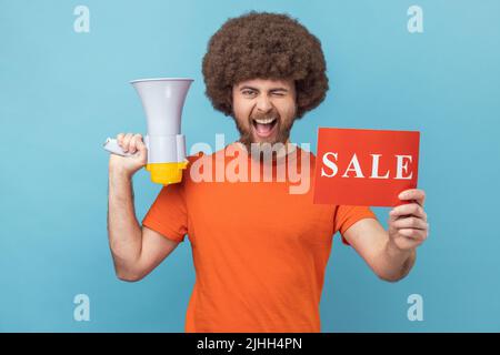 Porträt eines lustigen Mannes mit Afro-Frisur in orangefarbenem T-Shirt mit Verkaufsaufschrift und Megaphon, winkend zur Kamera, tolle Rabatte. Innenaufnahme des Studios isoliert auf blauem Hintergrund. Stockfoto