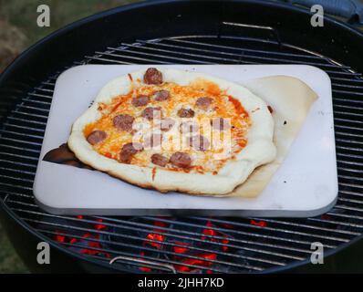 Eine hausgemachte Wurstpizza auf einem Pizzastein über einem Holzkohlegrill Stockfoto
