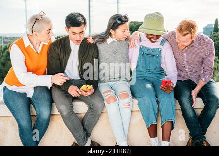 Fünf multirassische Freunde, die sich ein Video ansehen, mit zwei Smartphones, die lächelnd zusammen sitzen und Spaß haben. Stockfoto