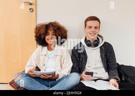 Zwei lächelnde Klassenkameraden blicken auf die Kamera. Fröhliche Studenten sitzen zusammen an der Wand mit Büchern. Stockfoto