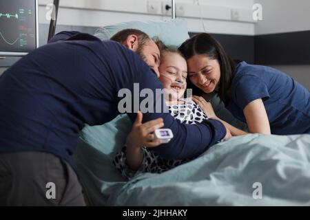 Fröhliche Mutter und Vater umarmen kranke Tochter sitzen im Patientenbett während der Behandlung. Glückliches krankes kleines Mädchen, umarmt von freudig lächelnden Eltern in der Kinderklinik. Stockfoto