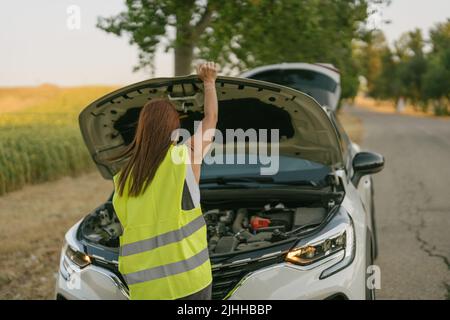 Rückansicht einer Frau mit einer reflektierenden Weste, die die Kapuze ihres Autos öffnet Stockfoto