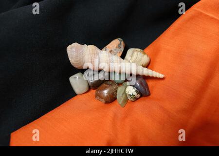 Eine Gruppe polierter Edelsteine auf schwarzen und orangen Stoffservietten mit einer Schneckenschale. Stockfoto