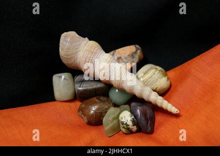 Eine Gruppe polierter Edelsteine auf schwarzen und orangen Stoffservietten mit einer Schneckenschale. Stockfoto