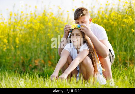 Der Bruder des Teenagers flechzt mehrfarbige Bänder in einem gelb-blauen Kranz mit wilden Blumen auf dem Kopf der fröhlich lächelnden Schwester auf der grünen Wiese A Stockfoto