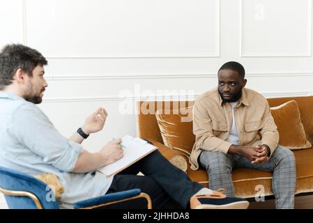 Ein schwarzer männlicher Patient, der sich einer Psychotherapiesitzung mit einem Berater in einer psychiatrischen Klinik unterziehen muss. Junger Mann mit emotionalen Problemen konsultiert die Profess Stockfoto